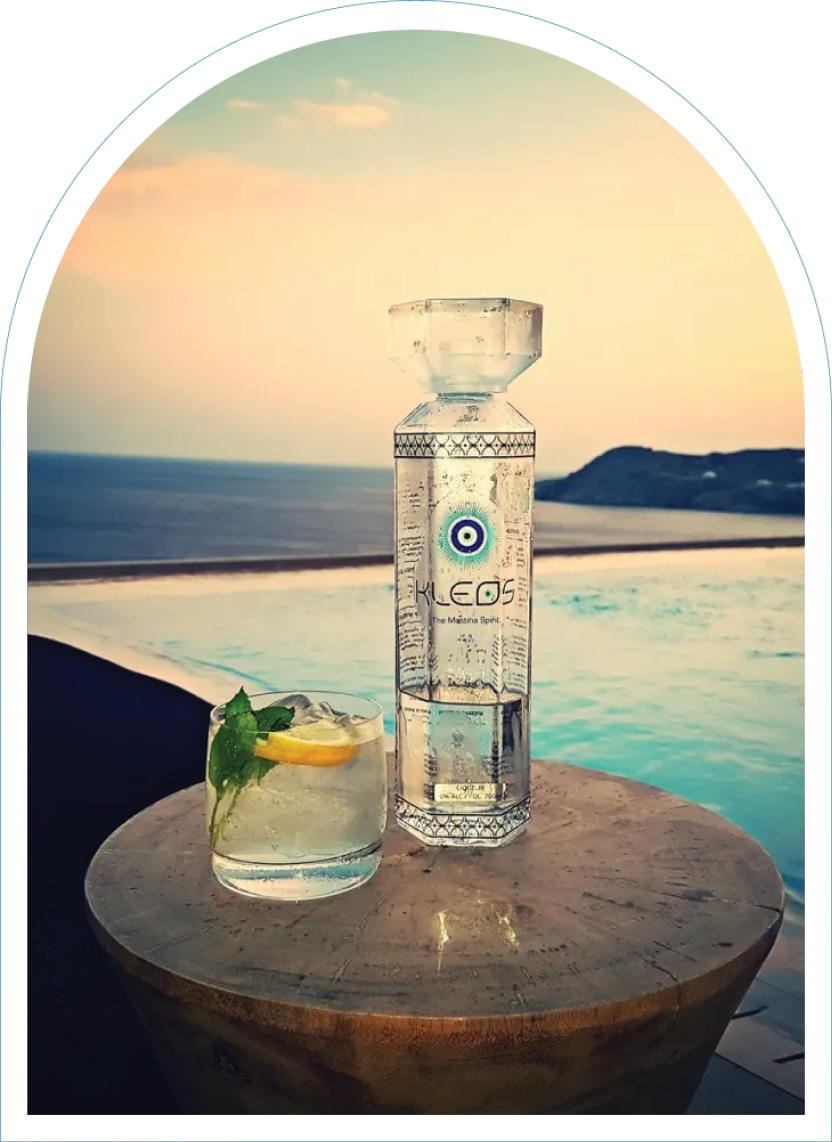 Bottle of KLEOS on a table in front of swimming pool at sunset. Mastiha tasting notes include lemon, cucumber, mint.