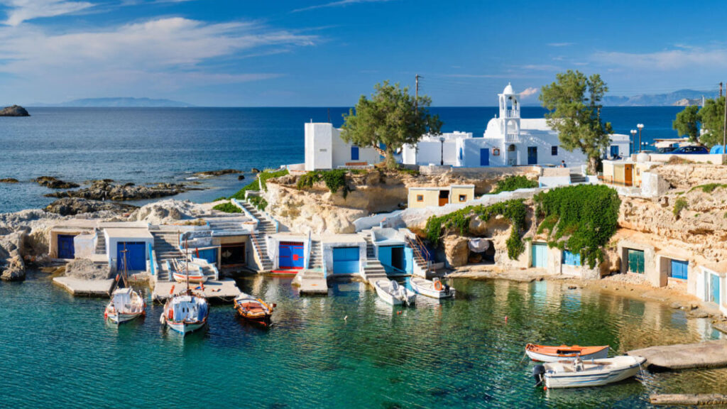 Small boats docked in a port with blue colored doors and the ocean in the background, on the island of Milos