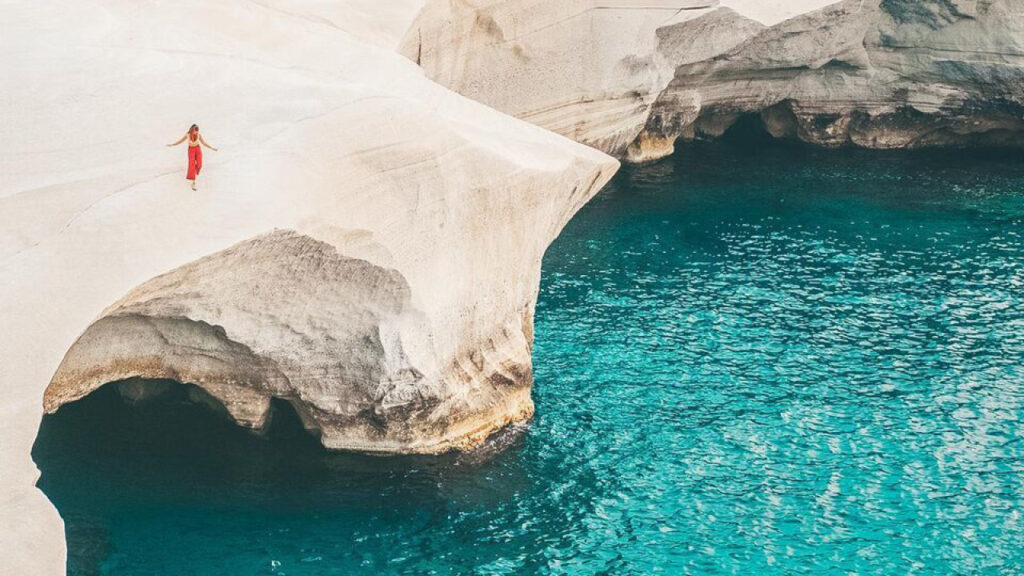 A person walking on top of a large cave opening, over the sea, on the island of Milos.