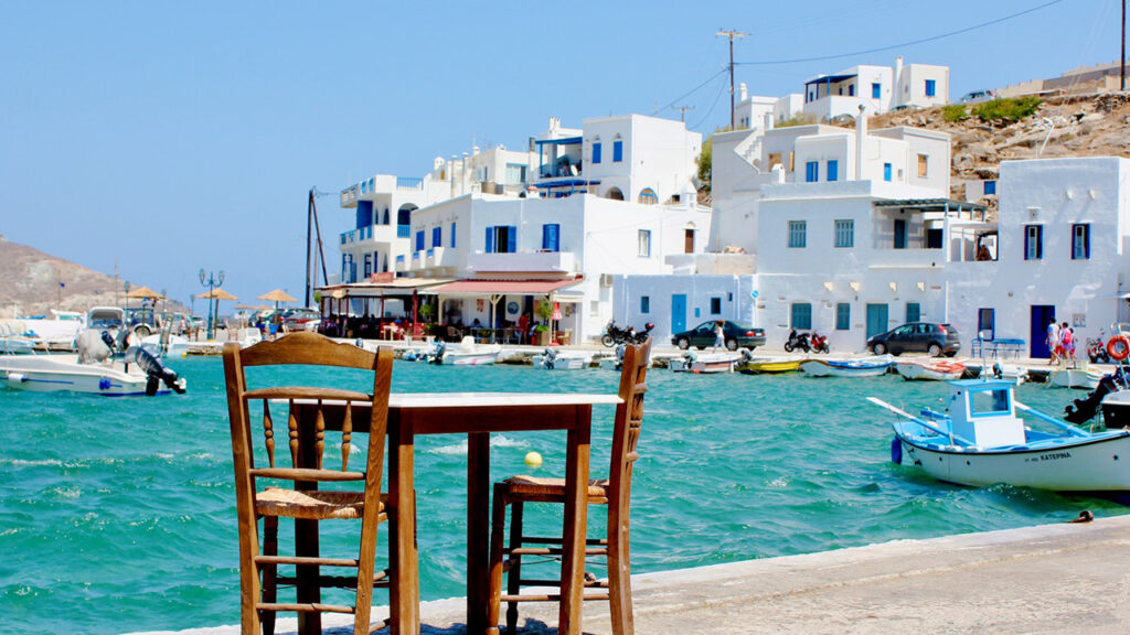 A table and two chairs close to the water in a harbor of Tinos, with some small boats and houses in the background.